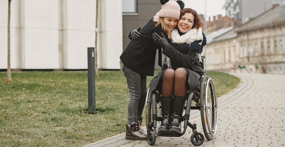   Mädchen mit Mütze und Frau in Rollstuhl umarmen sich und lächeln.